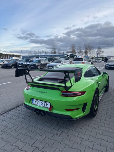A bright green Porsche 911 GT3 RS MR, bearing Polish registration plate W2 STF, is parked in a large outdoor paved lot in Wolica, Poland. The vehicle is positioned with its rear-right quarter facing the viewer, resting on grey interlocking concrete pavers. It features a prominent black rear wing with "MR Manthey" branding on the right upright. A red tow hook is affixed below the rear license plate, and two black exhaust pipes are visible centrally. The rear wheels are black with a neon green accent ring. The background reveals an expansive parking area filled with numerous parked and stationary vehicles, including sedans, SUVs, vans, and multiple large white passenger buses. Beyond the vehicles, a line of bare deciduous trees and evergreen conifers define the horizon. The sky is overcast with various shades of grey clouds. No discernible human activity or interactions are clearly visible in the immediate vicinity of the primary subject.