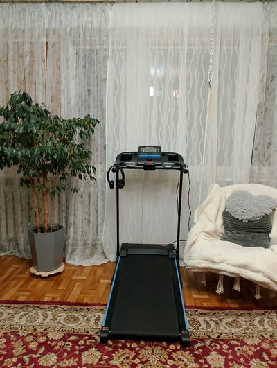 The image shows an indoor scene, likely a living room or exercise space, with a treadmill as the central focus. The treadmill is black with blue accents on the running belt and is positioned on a patterned red rug. To the left of the treadmill is a potted ficus tree in a hexagonal grey planter, which is elevated on a wooden base. In the background, sheer white curtains with a subtle floral pattern hang across a window. To the right of the treadmill, a plush, cream-colored armchair or beanbag chair is draped with a thick white blanket. A gray heart-shaped pillow with a ribbed texture rests on the blanket. A metal chain is suspended from the ceiling, connected to a braided rope that hangs down, suggesting the presence of a swing or hanging chair that is out of frame. The flooring is made of polished wooden planks arranged in a herringbone pattern. The overall impression is a home exercise setup combined with comfortable seating, suggesting a residential environment. There are no people visible in the image. The scene does not provide specific visual cues that would indicate its location in Kolno, Poland.