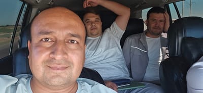 Three adult males are seated in the interior of a vehicle, likely a car, appearing to be in transit. The man in the foreground, facing the camera, is a close-up selfie. He has a shaved head, brown eyes, and is smiling. He is wearing a light blue t-shirt. To his right and slightly behind him, a second man is visible, also looking at the camera with a relaxed expression. He has dark hair and is wearing a light grey t-shirt. His left arm is raised and resting on the headrest of the seat behind him. Further to the right, a third man is seated, looking towards the camera with a neutral expression. He has dark hair and is wearing a light grey jacket over a white t-shirt. His right hand is resting on his head. The background shows the interior of the vehicle, including black leather seats and tinted windows. Through the windows, a landscape with dry vegetation and a road can be seen, suggesting an outdoor environment. The presence of the road and landscape points to the vehicle being on a journey, possibly outside of a dense urban area. This scene is captured in the city of Guzar, Uzbekistan.