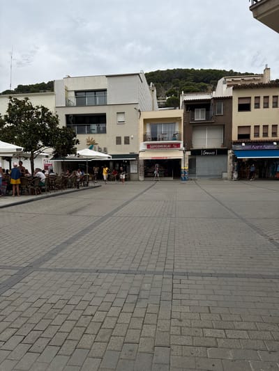 This image depicts a paved public square in L'estartit, Spain, under an overcast sky. The square's surface is composed of light grey rectangular pavers arranged in various patterns, with darker grey lines forming a grid.

Along the left side, an outdoor dining area is established, featuring approximately ten dark wicker-style tables and chairs positioned under several white market umbrellas. These are located in front of a multi-story building, the ground floor of which is occupied by "Alba," identifiable by a sign with a sun logo. A large tree with dark green foliage stands to the immediate left of this outdoor seating.

Approximately 15-20 individuals are present. A group is seated at the outdoor tables, engaged in dining or socializing. Several other individuals are walking across the paved square, including one person in a yellow top and dark shorts, and another in a white top and red shorts.

To the right of the "Alba" building, a sequence of commercial structures lines the square:
1.  A two-story building with a light yellow facade, housing a "SUPERMERCAT" on its ground floor. An A-frame sign and merchandise racks are displayed outside.
2.  A three-story building with a dark brown facade. Its ground floor features a closed metal roller shutter, displaying the name "Stelle."
3.  A multi-story, light-colored building on the far right, partially visible. Its ground floor hosts a shop named "Bali Kupu Kupu," identified by its blue awning and displayed clothing.

A densely forested hill, covered with dark green trees, rises directly behind the line of buildings, establishing the background environment. An antenna is visible extending above the leftmost building.