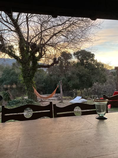 This image captures a peaceful outdoor scene, likely from a veranda or patio, looking out towards a garden. In the foreground, a wooden table is visible, with a clear glass on its surface. Beyond the table, a dark wooden bench with a circular cutout is positioned as a barrier.

The garden area features a large, ivy-covered tree with intricate, bare branches silhouetted against a soft, pastel-colored sky, suggesting either dawn or dusk. An orange hammock is suspended between two points in the garden, inviting relaxation. Several large green bushes and trees form a lush backdrop, with a hint of rolling hills or mountains in the distance. There is also a glimpse of red seating furniture further in the garden.

The environment appears serene and natural, with evidence of outdoor living and leisure. The overall atmosphere is tranquil and inviting. The location is identified as El Rodeo, Argentina.