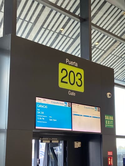This image depicts an airport gate area in the city of Tocumen, Panama, likely within Tocumen International Airport (PTY). The central feature is a dark grey gate structure labeled "Puerta 203" (Gate 203) with the number prominently displayed in black on a yellow-green background. Below this, two integrated digital screens provide flight information. The left screen shows a Copa Airlines flight CM 330 to CARACAS, with a scheduled departure of 12:06 PM on March 12, 2020, and a "Gate Open" status. The right screen displays "Upgrade List" for Puerta 203, also for Copa Airlines. Below these screens, an automatic glass door with a sensor bar above it is partially open, revealing a glimpse of an outdoor area or a parked aircraft's tail section with blue and white stripes. A small, white dome-shaped security camera is mounted on the dark grey column above the right screen. To the right of the screens, a green "SALIDA EXIT" sign is visible, and below it, a red fire extinguisher with a "DESLICE" (Slide) instruction is mounted on the wall. The background consists of large windows revealing a patterned ceiling structure or sun louvers, indicating a bright, modern airport terminal environment. No individuals are present in the frame.