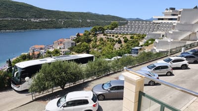 This is a high-angle, daylight, wide shot of a coastal city in Neum, Bosnia and Herzegovina. The scene is dominated by a large, white tour bus parked on a paved road. Several cars are also parked on the road, including a white SUV, a silver hatchback, and a gray sedan. In the background, there is a large, multi-story white hotel with a distinctive stepped facade, set against a backdrop of green, tree-covered hills and a calm, blue bay. Buildings with orange-tiled roofs are visible closer to the water. The day is clear with a bright blue sky. There are no people clearly visible in the image.
