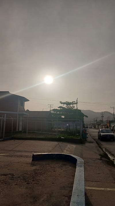 The image shows an outdoor scene in what appears to be a street, likely taken in Barcelona, Venezuela. The sun is high in the sky, causing a glare. On the right side of the image is a concrete curb with a painted blue edge. A paved road surface with lane markings is visible. On the other side of the road, there is a fence, trees, and some buildings. A dark-colored car is parked on the side of the street. The background suggests a townscape with buildings and a mountain or elevated land in the distance. The overall light condition suggests it is daytime.