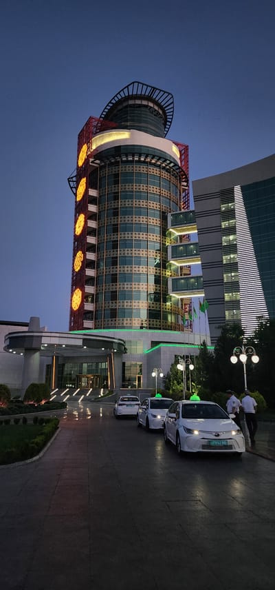 The image depicts a contemporary building with a curved, glass facade illuminated by warm lights. The building is adorned with decorative red metalwork and circular yellow lights at its base. To the right of the building, several white vehicles are parked in a line on a paved driveway. Two individuals, dressed in white shirts and dark pants, stand near the vehicles, appearing to be interacting. The scene is set at dusk, with a deep blue sky overhead. Lush green trees and manicured bushes surround the driveway, adding to the sophisticated ambiance of the location. The overall impression is one of a well-maintained and modern establishment. The license plate on the front car reads "A 7952 BB".