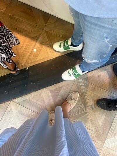 The image shows the feet and lower legs of at least four individuals standing on a parquet floor, separated by a black threshold. From left to right, the first person is wearing a black sandal with a white strap and a black and white patterned garment, likely a dress or skirt. Their foot is positioned on the left side of the frame. Next to them, a person wearing distressed light-wash denim jeans and white sneakers with green stripes stands with their feet on the right side of the black threshold. Their sneakers have prominent white laces and a white sole with triangular studs. Further to the right and lower in the frame, another individual is wearing light brown loafers with tassels. Their foot is placed on the lower right quadrant of the image. To the right of the loafers, a person is wearing black athletic shoes with white accents. The floor is a light-colored parquet with a geometric pattern. The lighting suggests an indoor environment, possibly a shop or a public space. The image does not provide specific details to determine the exact location within Chevilly-Larue, France, or any particular activity beyond standing.
