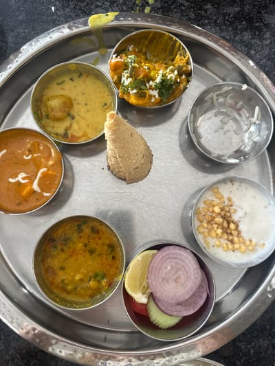 A close-up, high-angle shot shows a traditional Indian thali served on a round, silver platter. The platter contains seven small metal bowls and a single papadum. Starting from the top and moving clockwise, the bowls contain: a creamy yellow curry with what appears to be a fried fritter; a vegetable curry with white crumbled cheese and cilantro garnish; a small amount of white rice; a bowl of yogurt with small, fried chickpea flour balls and a sprinkle of spice; a small bowl of salad with sliced red onion, cucumber, and lemon; a thick, orange curry with paneer or tofu and a white cream drizzle; and a yellowish-orange lentil dal. The papadum is placed in the center of the platter. The background is a dark, speckled surface, likely a table. The overall impression is of a neatly arranged, appetizing meal. The location is identified as Dhani Gaiskan, India.