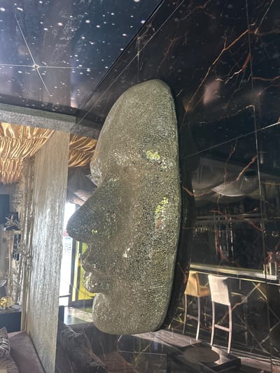 The image captures an indoor scene with a large, translucent mask sculpture as the central subject. The mask is made of a textured, possibly glass or resin material, with a mosaic-like surface that reflects light. It is positioned prominently in the foreground, appearing to be suspended or mounted.
The background reveals a richly decorated interior. To the left, there are layers of shimmering gold fabric curtains and textured wall coverings, suggesting an opulent or theatrical setting. Further left, a glimpse of furniture and decorative elements, possibly plants or sculptures, is visible.
The right side of the image shows a dark, reflective wall with a marbled pattern featuring prominent red and orange veins. This wall reflects a bar area with light-colored stools. The ceiling above has a dark, starry pattern, possibly a custom design or a printed material, which further enhances the atmospheric and potentially surreal quality of the space.
The overall impression is of an interior designed for visual impact, likely a commercial space like a lounge, club, or art installation. The scene is static, with no people visible, and the focus is entirely on the decorative elements and the striking mask sculpture. The location of Brca, Montenegro is not evident from the visual cues in the image.