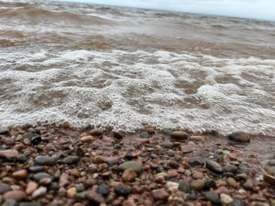 A low-angle, close-up shot captures the edge of a body of water meeting a rocky shore. The water is a muted reddish-brown, with white foam and bubbles creating a textured line where it washes over the pebbles. The pebbles themselves are a mix of sizes and colors, predominantly reddish-brown and gray, with some larger, smoother stones visible. The background shows the rippling surface of the water extending to the horizon, meeting a hazy, overcast sky. There are no people or animals present in the scene, and the focus is on the natural elements of the shore and water. This scene could be found along the coast of the city of Kostovaty, Russia, where similar geological features and water bodies are common.