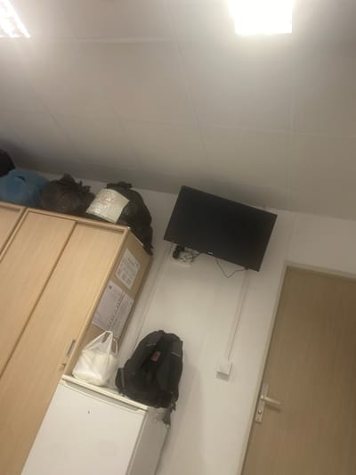 The image depicts a cluttered room, possibly a dormitory or storage area. A wooden wardrobe is visible on the left, with items stacked on top, including several black trash bags and a blue bag. A white refrigerator sits below the wardrobe, with a white plastic bag and a black backpack placed on top of it. A flat-screen television is mounted on the wall to the right, with wires connected to a white electrical outlet below. A light fixture is visible in the ceiling. A closed wooden door is to the far right of the image. The room appears to be located in Қосшағыл ауылдық округі, Kazakhstan, based on the provided information.