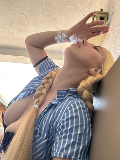 A blonde woman, seen from a low angle, is smoking a cigarette outdoors. She is wearing a blue and white striped shirt. Her blonde hair is braided and falls over her shoulder. The background shows a textured wall and a wooden birdhouse or electrical box. Sunlight suggests it is daytime. The woman appears to be exhaling smoke. The overall setting suggests a casual, possibly residential, outdoor environment.