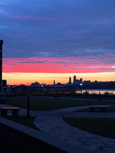This is a vertical, eye-level, outdoor photograph taken at dusk in Edgewater, United States of America. The image depicts a cityscape on the horizon, silhouetted against a vibrant sky. The sky is a dramatic gradient of deep blue at the top, transitioning to streaks of bright pink and orange lower down, indicative of a sunset or sunrise. Wispy clouds are visible, reflecting the sky's colors.

In the foreground, a paved walkway with a textured, stone-like pattern winds through a grassy area. Several stone benches are strategically placed along the walkway. A tall, dark pole with a light fixture is visible in the middle ground. To the left, a dark, multi-story building with windows is partially visible, dominating the left edge of the frame.

Beyond the grassy area and walkway, a body of water separates the foreground from the distant cityscape. The city skyline is a dark silhouette, featuring various buildings of different heights, including some taller, more prominent structures. A long, low building with a pier extending into the water is also visible in the middle distance. The overall scene conveys a sense of calm and natural beauty, juxtaposed with the urban landscape. There are no people or animals visible in the frame.