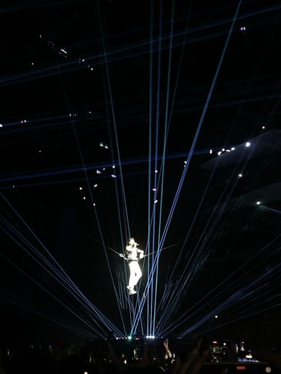 A person suspended mid-air in a white suit, attached to ropes, is the focal point of the image. The background is dark and pierced by numerous blue laser beams that converge on the right side. There are also small floating, illuminated particles, possibly confetti or dust. A small portion of the crowd is visible on the right, holding up phones. The overall impression is one of a theatrical or performance event.