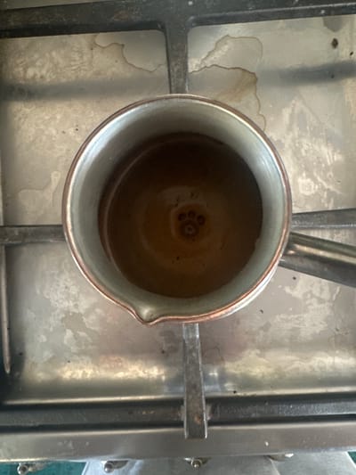 A top-down, close-up shot shows a copper cezve filled with dark brown liquid, likely coffee, resting on a metal stovetop. The liquid has a thick crema on top, with a distinct four-lobed pattern resembling a paw print in the center, likely formed by the bubbling of the coffee. The stovetop itself appears to be of a brushed metal material, with some dark stains and smudges visible, suggesting it has been used recently. The cezve has a rounded body and a long, thin handle extending to the right, also made of metal. The lighting is overhead, casting subtle shadows and highlighting the textures of the metal surfaces. The location of this scene within Сарымолдаев ауылдық округі, Kazakhstan, is not directly discernible from the visual information provided in the image itself.