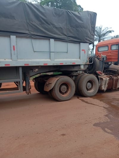 The image shows the rear of a large orange semi-trailer truck with a grey trailer attached. The truck has a spare tire mounted behind the cab. The trailer is covered with a large grey tarp. The truck and trailer are parked on a dirt road. There is some water on the road, suggesting it may have recently rained. In the background, there are trees and a palm tree. The words "ANAMCO" are visible on the side of the truck's cab. This scene is located in Imilike, Nigeria.