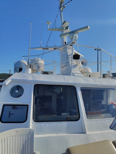 The image shows the upper deck and superstructure of a white boat, possibly a ferry or a research vessel, under a clear blue sky. The boat has several antennas, radar equipment, and a searchlight mounted on a central mast. Large windows with horizontal blinds are visible on the side of the superstructure. A white plastic chair is in the foreground, and part of a padded seat is visible on the right. The background shows a hilly landscape with some vegetation and a power line pole. The boat is docked, as indicated by the clear blue water and the presence of shore facilities in the distance. The location is identified as Municipal Unit of Pelasgia, Greece.