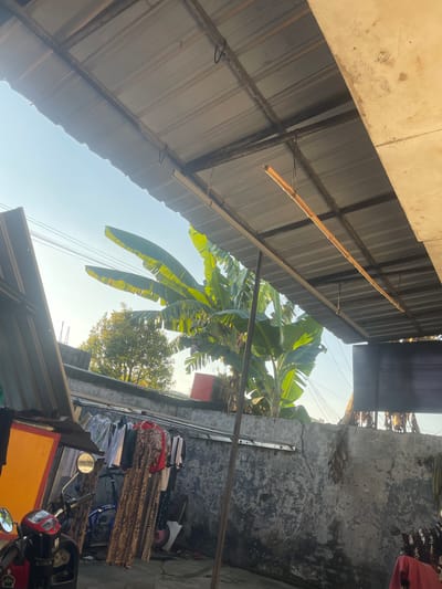 The image depicts an outdoor scene under a corrugated metal roof supported by metal beams. A clothesline is strung across the area, with various garments hanging on it, including dresses and t-shirts. In the foreground, a red and black motorcycle is parked, with its mirrors visible. To the left, an orange structure is partially visible. Behind the clothesline, a grey, weathered concrete wall runs along the background. Banana trees with large green leaves are prominent, and a smaller, leafy tree is visible further in the distance. The sky is clear and bright blue. The scene suggests a residential area, possibly a backyard or an alleyway, in Anggaswangi, Indonesia.