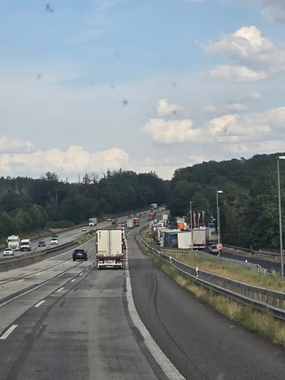 The image shows a traffic jam on a multi-lane highway surrounded by dense forest. The highway appears to be the Autobahn A3, as indicated by the location context of Heiligenroth, Germany. Numerous trucks and cars are stationary or moving slowly. The sky is partly cloudy with a blue hue. In the foreground, the road surface shows tire marks. The scene is captured from a moving vehicle, likely another car on the highway.