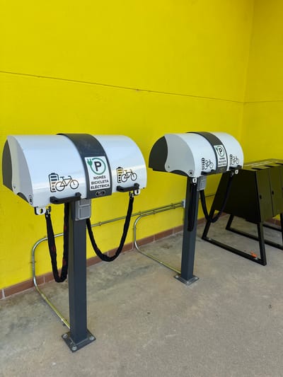 The image shows two electric bicycle charging stations positioned in front of a bright yellow textured wall. Each station has a white, curved housing with a black stripe around the top. The housing features icons of a bicycle and a battery, along with text that appears to be in Catalan: "NOMÉS BICICLETA ELÈCTRICA" and "P" inside a green circle, indicating parking for electric bicycles. A black chain hangs from each station, presumably for securing the bicycles. To the right of the charging stations is a black metal rack with multiple compartments. The ground is concrete, and metal conduits run along the wall at the base. The scene appears to be an outdoor public space, possibly a street or a park, in the city of Vidreres, Spain, given the Catalan text. No people are visible in the image.