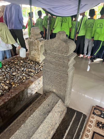 In Beuringen, Indonesia, an outdoor scene is captured, likely a historical or religious site. The foreground features a stone tomb with steps leading up to it, constructed from rough-hewn granite. Beside the tomb is a grave enclosure filled with pebbles and a white plastic jug with a blue cap. In the mid-ground, a group of children, some wearing light green shirts with the text "MADRASAH IBTIDAIYAH NEGERI 10 ACEH UTARA" and black skullcaps, stand under a white canopy. Some of the children are wearing black pants, while others are in light grey pants. To the left of the tomb, an adult in a light blue long-sleeved shirt and dark pants stands. The ground is a tiled floor that reflects the light. A small rug with a patterned design is visible on the right side of the frame.
