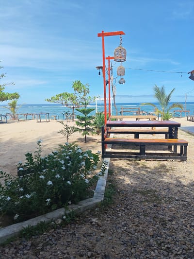 The scene is outdoors near the ocean. It appears to be a coastal area, possibly a beach or a restaurant located near the beach. The location is in Monikeun, Indonesia.

Visible in the foreground are white flowering plants next to a gray concrete border, and a gravel surface. In the mid-ground there is a wooden table with attached benches. Suspended from an orange metal structure are decorative birdcages. Beyond the table, there is sand, sparse vegetation (including palm trees and small trees), and other tables with benches visible in the distance. The background shows the ocean with a clear blue sky with a few clouds. There are no people visible in the image.