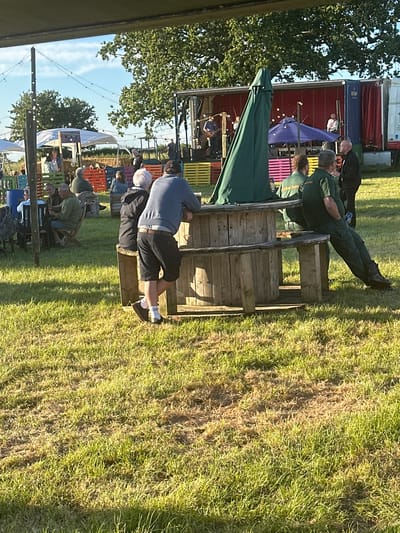 The image depicts an outdoor event with a stage and a seated audience. A band is performing on the stage, which is set up on a truck. Several people are seated in the foreground, facing the stage. Some are sitting at wooden spool tables and benches. There is a large green umbrella in the foreground, partially obscuring the view. The background shows trees, a blue sky with clouds, and some tents and structures. The overall impression is of a daytime outdoor gathering or festival. No specific location in the city of Gloucester, United Kingdom, is identifiable from the visual information in the image.