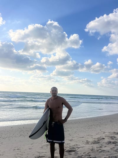 The image depicts a male subject standing on the sandy beach of Beit Yanai, Israel. The man, appearing to be in his twenties or thirties, is shirtless and wearing dark swim shorts with a white horizontal stripe near the hem. He has short dark hair, a light-colored necklace, and bracelets on his left wrist. He holds a light-colored surfboard with black trim and the text "AFSL" or "AFSL" visible on its side under his left arm. A black leash or strap is attached to the surfboard. His right hand is positioned on his hip, and he is looking directly at the camera with a slight smile.
Behind the man, the Mediterranean Sea exhibits moderate waves breaking towards the shore. Several indistinct figures, likely other individuals in the water, are visible in the distance. The sky above is partially covered with scattered cumulus clouds against a blue background, suggesting either late afternoon or early morning light. The foreground consists of wet and dry sand with visible footprints.