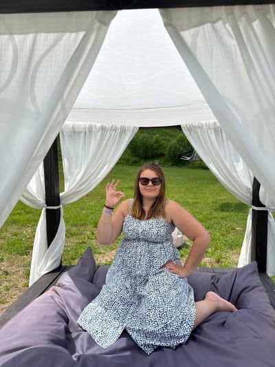 A young woman with brown hair, wearing sunglasses and a patterned dress, sits on a large grey cushion inside a white gazebo. She is making an "okay" sign with her right hand and has her left hand on her hip. The gazebo has white curtains that drape down on the sides, framing the scene. Outside the gazebo, a grassy area with trees is visible. The location is identified as Nechkino, Russia.