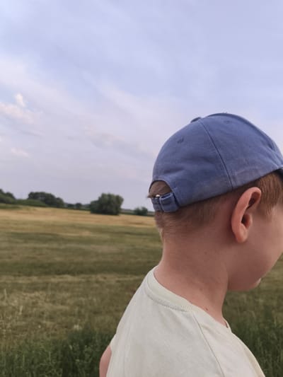 The media file is a close-up, side-view shot of the back of a young person's head and shoulders. They are wearing a light blue baseball cap and a light-colored t-shirt. The person has short, light brown hair visible beneath the cap, and their right ear is clearly visible.
The background consists of a wide, grassy field with some trees dotted in the distance, suggesting a rural or park-like setting. The sky is overcast with light clouds, indicating it might be late afternoon or early evening, and the weather appears calm.
The focus is on the person, with the background slightly blurred, creating a sense of depth. The lighting is soft, consistent with an overcast sky. There are no visible signs of a specific activity or event, other than the person appearing to be outdoors and possibly looking towards something in the distance. The overall mood is serene and peaceful. No text is visible in the image.
Given the location context of Stanisławowo, Poland, the scene likely depicts a moment of quiet contemplation or enjoyment of nature in a Polish landscape.