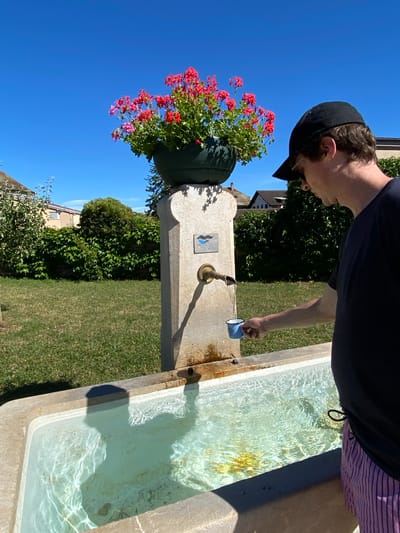 In Boudry, Switzerland, a person wearing a black baseball cap and a dark t-shirt is filling a small blue mug with water from a stone fountain. The fountain has a brass spigot from which water is flowing into the mug and a larger basin below. The fountain is adorned with a pot of vibrant pink geraniums. The scene is set outdoors on a sunny day, with a clear blue sky and green grass in the background, along with some trees and buildings.