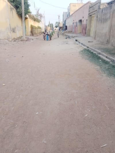 A long, dirt road stretches through a village, flanked by buildings of varying heights and construction styles. A drainage ditch lines the right side of the road, partially filled with grass. In the distance, a group of people walks towards the camera, carrying items on their heads. The sky is hazy, creating a muted color palette. The overall scene depicts a simple, rural life.
