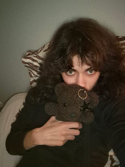 A person with long, dark, curly hair and light-colored eyes is lying down, holding a small, dark brown teddy bear close to their chest. The teddy bear has a black star and a golden ring attached to it. The person is wearing a dark-colored long-sleeved shirt. They are lying on a bed with a zebra-striped pillow or blanket near their head, and a white sheet or blanket underneath them. The background appears to be a plain wall or surface. The image is rotated slightly to the left.
