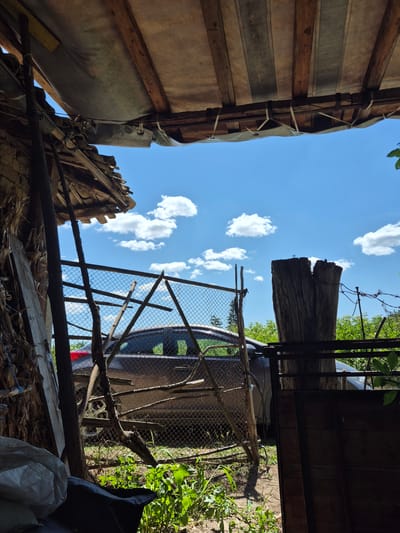 The image depicts an outdoor scene under a rustic, makeshift roof structure in Sadina, Bulgaria. The roof is made of wooden beams and covered with a tarpaulin, with a section of corrugated material visible. Beneath the roof, a chain-link fence encloses an area where a dark-colored car is parked. The car appears to be a hatchback. The fence is supported by wooden posts and branches. To the left of the fence, there are stacks of dried plant material, possibly corn stalks, and some large white sacks. To the right of the fence, a large, weathered wooden post stands, with barbed wire attached to it. Lush green vegetation is visible behind the fence and around the wooden post. The sky above is blue with scattered white clouds. The overall impression is one of a rural or semi-rural setting, likely a yard or storage area. There are no people visible in the image.