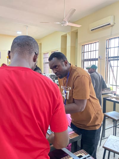 Three men are indoors, possibly in a classroom in Afaka, Nigeria. One man is wearing a red shirt and is closest to the camera, with only the back of his head and upper back visible. Another man is wearing a tan t-shirt with writing on it and a blue lanyard around his neck, leaning forward, possibly looking at a document or object on a table. The third man is wearing a gray shirt and a green and white baseball cap, also leaning forward and appearing to look at something on the table. The room has yellow walls, windows with bars, a ceiling fan, and an air conditioning unit. There are tables and chairs in the background.