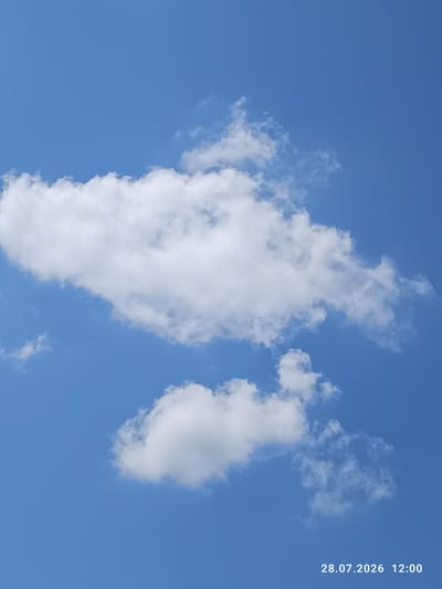 The image shows a bright blue sky with scattered white clouds. In the bottom right corner, there is a timestamp indicating "28.07.2026 12:00". There are no people, animals, buildings, or other recognizable objects in the scene. The image is a straightforward photograph of the sky.