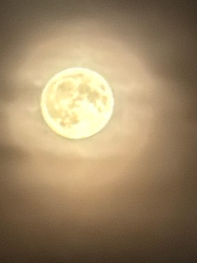 The image shows a full moon with a hazy glow around it. The moon is a bright, yellowish-white sphere with some visible craters or surface features. It is set against a dark, out-of-focus background that appears to be a night sky with some atmospheric haze or light pollution. The overall impression is that of a celestial body observed from Earth. The image does not depict any specific location in the city of Kamianka-Buzka, Ukraine, nor does it show any people, objects, actions, or interactions.