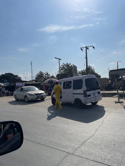 The image depicts a daytime street scene in Ibeju, Nigeria, under a bright, clear sky with high sun and minimal cloud cover. The environment includes a paved road with visible cracks, various single-story buildings and market stalls with awnings in the background, trees, and utility infrastructure.
**People:** A sole individual, dressed in a bright yellow two-piece uniform, walks from the left to the right side of the frame across the roadway, slightly behind a white van. This individual holds a dark, circular object, possibly a tire or a bag, in their left hand. Other indistinct figures are visible near the market stalls in the background. A reflection of a person's arm is visible in the bottom-left side mirror of the vehicle from which the photograph was taken.
**Subjects/Objects:**
* **Vehicles:** A white commercial minivan or passenger van, exhibiting significant wear and damage to its rear bodywork, is positioned facing away from the camera, slightly off-center to the right. A silver four-door sedan (identified as a Toyota Camry) is positioned on the left side of the frame, facing slightly right, displaying a Nigerian license plate. Several motorcycles or bicycles are partially visible behind the sedan, near the market stalls.
* **Infrastructure:** Multiple wooden utility poles with overhead power lines are visible, along with at least two solar-powered streetlights. A tall, lattice-style telecommunications tower is present in the distant left background.
* **Market Stalls:** Several open-air market stalls with colorful umbrellas and various goods are discernible in the background, suggesting commercial activity.
**Actions/Interactions:** The individual in yellow is actively walking across the road. The vehicles appear to be stationary or moving slowly within traffic. The overall scene indicates general pedestrian and vehicular activity characteristic of a local thoroughfare or market area. Strong sunlight casts long, distinct shadows from the vehicles and the walking individual across the road surface.