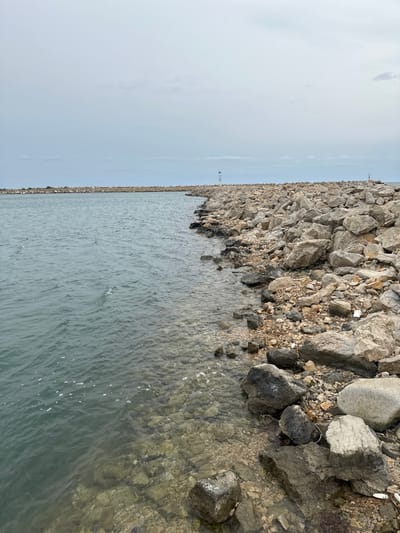 The image displays a coastal scene in Limenas Avdiron, Greece, characterized by a breakwater and calm water under an overcast sky. The upper two-thirds of the frame are occupied by a light grey sky with minimal cloud definition. The lower portion features a body of water on the left, exhibiting a greenish-blue hue with visible surface ripples, indicating minimal current. Closer to the right, the water becomes shallower and clearer, revealing a rocky seabed composed of varying sizes of light-colored stones and sediment.
A prominent man-made breakwater, constructed from irregularly shaped grey and brown rocks of diverse sizes, extends from the mid-right foreground into the background, curving slightly towards the left. This structure separates the calmer inner water from what appears to be the open sea beyond its farthest extent. Two white navigation markers are positioned along the breakwater. The more discernible marker is a slender, white cylindrical pole with a black conical top, located approximately midway along the visible length of the breakwater. A second, smaller, and less defined white structure is visible further down the breakwater towards the distant horizon on the right. No individuals or animate subjects are present in the scene. Small fragments of debris, consistent with plastic, are scattered among the rocks on the breakwater in the foreground.