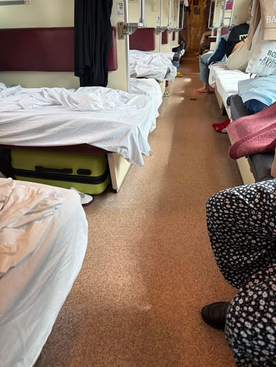 A high-angle shot captures a row of train berths, likely in a compartment, filled with luggage and people. The scene is somewhat cluttered, with white bedding covering most surfaces, including a large suitcase on the floor. People are visible in various states of repose or travel, some with legs extended or feet showing. One person is wearing a black floral dress and dark shoes, another is wearing red sneakers. A person in the foreground is partially visible, draped in white fabric, with a hint of a light blue shirt beneath. A green suitcase is neatly placed under a shelf, and a black garment is folded on a maroon seat. The flooring appears to be a cork-like material. The overall impression is one of travel and the temporary arrangement of personal belongings in a confined space. The lighting suggests it could be daytime, with natural light filtering in, creating shadows. The image is oriented vertically, making the train's interior appear elongated. No specific emotional states are discernible from the visible subjects. There are some labels on the metal fixtures, including "12 11" and "05.17.18."