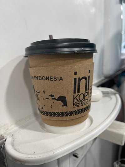 A close-up, eye-level shot shows a paper coffee cup with a black lid and a brown sleeve. The sleeve has black text and a silhouette of Indonesia. The text reads "KOPI INDONESIA" and "ini KOPI KERETA." The cup is sitting on a white tray, which is mounted on a white surface that appears to be the interior of a vehicle, possibly a train, given the text on the cup. The background is blurred and white. The location is not specified in the image or implied.