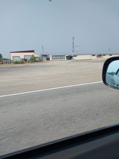 The image shows a street scene in Novotyryshkino, Russia. In the background, there are several industrial buildings with beige walls and corrugated metal roofs. One building has a red roof. Two tall communication towers are visible in the distance. In the foreground, a paved road with a white line marking the lane is visible. A side mirror of a car is in the lower right corner of the frame, reflecting a blurred image of the road and landscape. The sky is clear and pale blue. There are no people visible in the image. The scene appears to be an industrial or commercial area on the outskirts of the city.