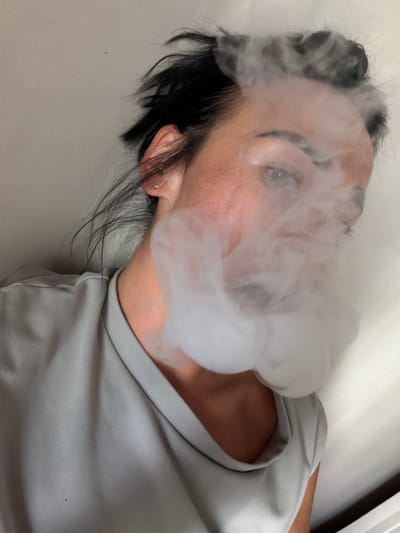 A woman is captured from a high angle, her face partially obscured by a cloud of white vapor, obscuring her mouth and nose. Her dark hair is styled in an updo, with strands falling loosely around her face and neck. She is wearing a light gray t-shirt. A small, sparkling stud earring is visible in her right ear. Her skin has a light complexion with scattered freckles. The background is a plain, light-colored wall, suggesting an indoor setting. There are no discernible landmarks or elements that would indicate a specific location within Koltushi, Russia.