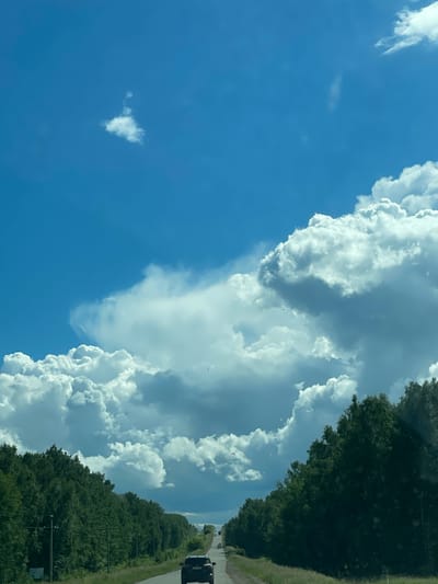 The image depicts a road passing through a forest with a car driving on it. The sky is filled with dramatic clouds. There is no indication of the location being the city of Лобаново, Kazakhstan in the image itself. The image contains one visible car, which is black and appears to be an SUV. There are no people visible in the image. The environment is a roadside with a dense forest on either side of the road. The road is asphalt and appears to be a two-lane highway. The time of day appears to be daytime, with bright sunlight filtering through the clouds. There are some telephone poles visible along the side of the road.