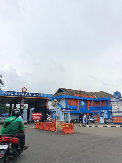 The image depicts the entrance to Sulaiman Air Force Base. A sign overhead indicates the base name, while a blue and orange building, labeled "RSAU Normant Lubis", stands to the right. Traffic barriers and a warning sign block the entrance. A motorcyclist in a green jacket rides towards the camera. The sky is overcast, and other motorcycles and people can be seen near the building.