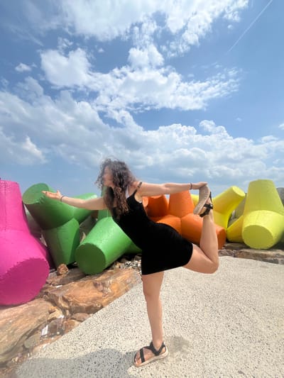 A woman with curly brown hair is performing a yoga pose, specifically Natarajasana (Lord of the Dance Pose), outdoors. She is wearing a black dress and sandals. Her left leg is bent, with her foot raised behind her and held by her left hand. Her right arm is extended forward. The background features a row of large, colorful concrete tetrapods, painted in bright pink, green, orange, and yellow. The ground is a textured paved surface. Behind the tetrapods, there is a rocky embankment. The sky above is blue with scattered white clouds, and a faint contrail from an aircraft is visible. The scene appears to be located in Phú Thủy, Vietnam, likely along a coastline or riverbank, given the presence of tetrapods which are often used for coastal defense.