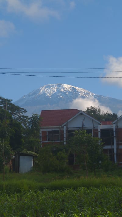 The image shows a mountain partially obscured by clouds, situated behind a building with a red roof in Longuo 'A', Tanzania. Power lines are visible in the blue sky. Dense green foliage and a small shed are in the foreground.