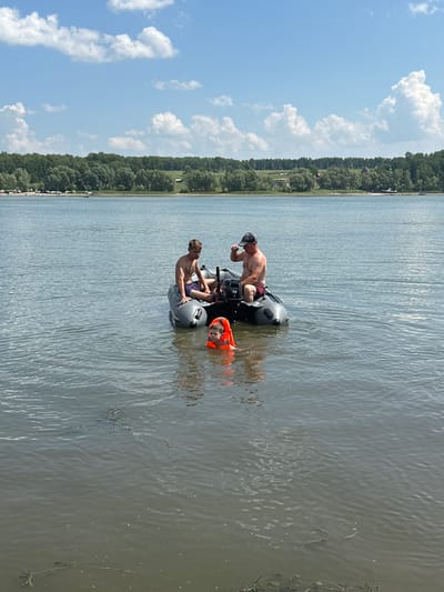 In the city of Pravda, Russia, on a calm, overcast day, three individuals are present on a body of water. A man wearing a black cap and red shorts is seated in a gray inflatable boat, holding a beverage. Beside him, another man in purple shorts sits in the same boat. In the water, a young child wearing an orange life vest is visible, partially submerged. The background features a dense line of green trees and a bright blue sky with scattered clouds. The water appears murky with some debris floating on the surface.