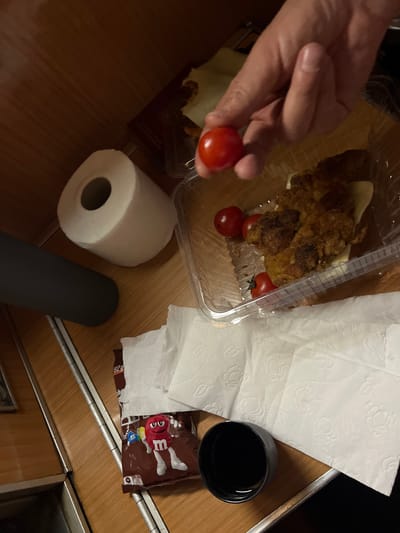 A top-down shot reveals a casual dining setup on a wooden surface, possibly a table. A hand holds a small, red tomato, apparently removed from a clear plastic container that also holds a fried food item topped with cheese and several other tomatoes. Next to the container are several white paper napkins, a roll of toilet paper, and a dark grey cylindrical object. A dark liquid in a black cup sits nearby, along with a package of M&M's. The items suggest a meal or snack situation, but there are no clues present to suggest any information regarding Lopate, Montenegro.