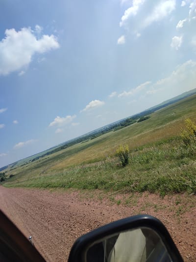 The image depicts a rural landscape with a dirt road in the foreground and a vast, rolling field stretching into the distance. The field is covered in a mix of green and golden grasses, with scattered wildflowers and small bushes. A line of trees borders the field in the middle ground, and beyond that, the land continues to undulate under a bright blue sky dotted with white clouds. A car's side mirror is visible in the lower right corner of the frame, reflecting a portion of the sky and some light-colored objects, likely the interior of the car. The scene suggests a sunny day in a countryside setting. There are no identifiable people or specific structures visible in the image. The location is identified as Bakhmut, Russia.