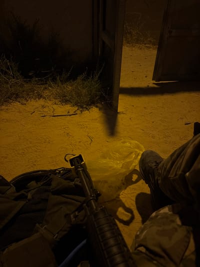 This media file captures a nighttime scene in the Eshkol Regional Council, Israel. The dimly lit ground appears to be sand or dirt, marked with scattered debris and what seem to be faint footprints. To the left, a patch of dry, wispy grass grows against a dark background, possibly indicating the edge of a cultivated area or a natural landscape.
In the foreground, the lower half of a person is visible, wearing dark pants and a dark-colored shoe. A bright yellow plastic bag lies on the ground near their foot. To the right, a portion of a rifle is prominently displayed, resting on what appears to be a dark backpack. The rifle has a distinctive yellow muzzle cap or marking. The overall lighting suggests artificial illumination, likely from a nearby lamp or streetlight, casting long shadows on the ground and creating a somber atmosphere.
The scene is devoid of any explicit action, suggesting a moment of pause or stillness. The presence of the rifle and the military-style clothing implies a security or operational context, possibly related to the location's proximity to border areas. No text is visible in the image. The time of day is clearly night, and there are no immediate cues about the weather, though the dry ground might suggest arid conditions.